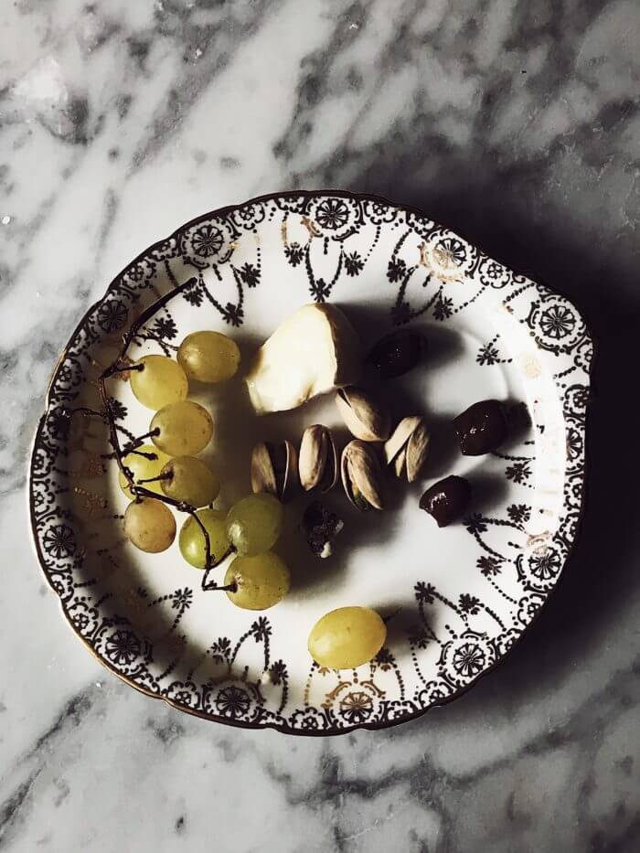 a slice of cheese, grapes and pistachios on a white and golden fine china plate