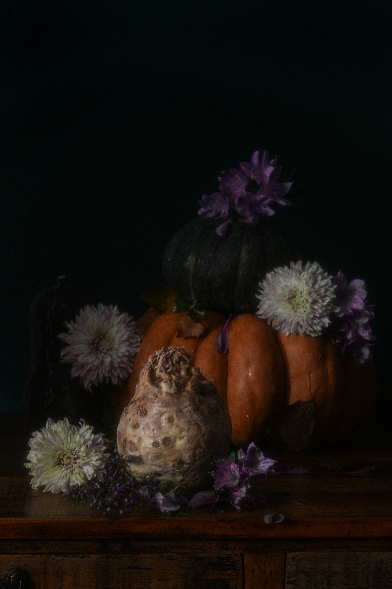 Dark autumn still life with orange and green pumpkins, a knobbly celeriac and purple and pale flowers arranged on an antique wooden surface.